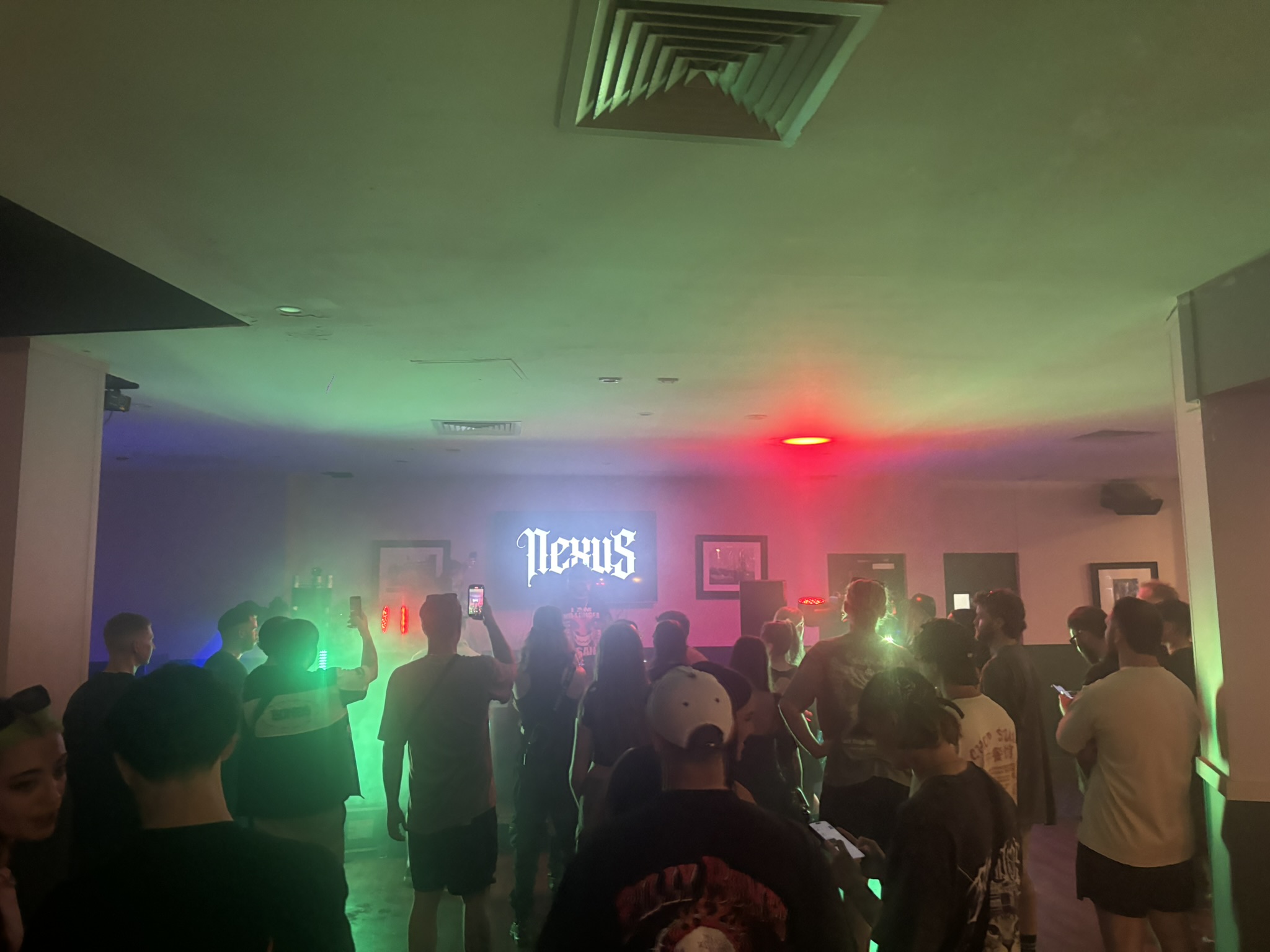 Crowd on the dance floor under green and red haze with the Nexus screen lit up behind